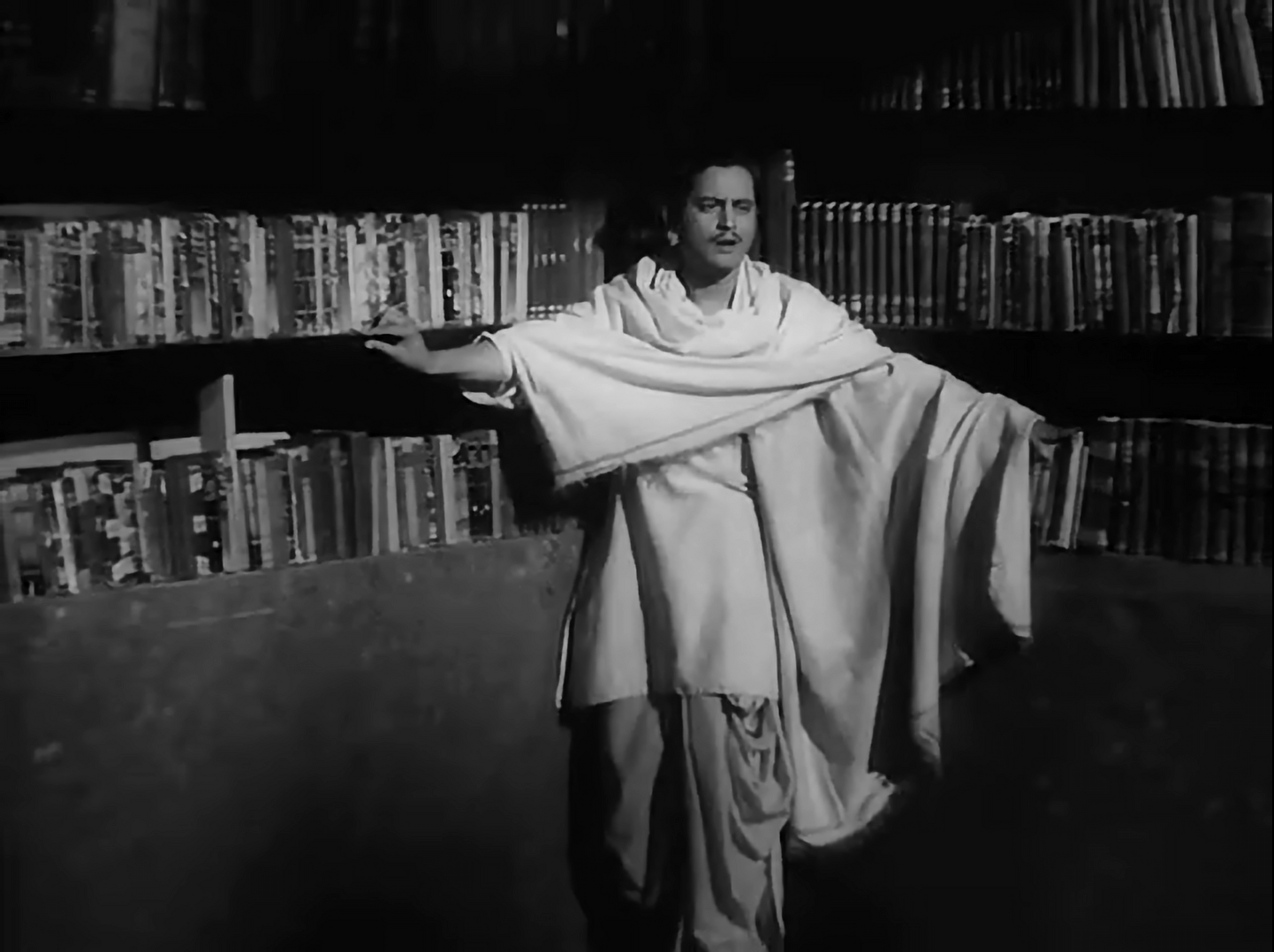 Guru Dutt stands with arms stretched against the bookshelf